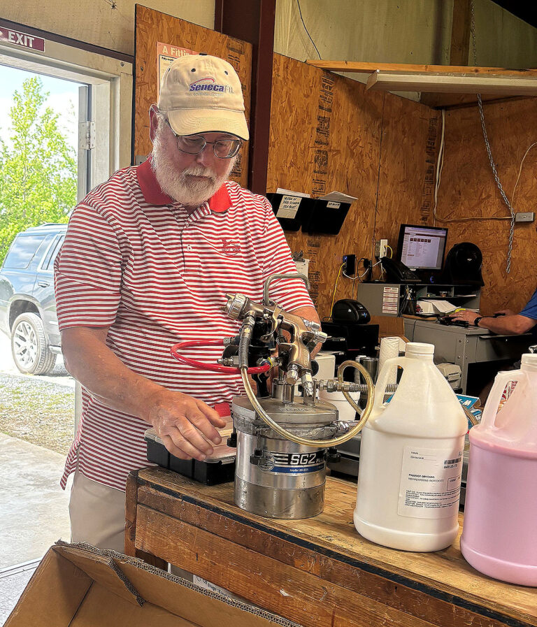 While standing at a workbench with a spray gun, pressure pot and bottles of glue, a technician explains how low pressure spraying of industrial adhesive works for precision bonding applications.