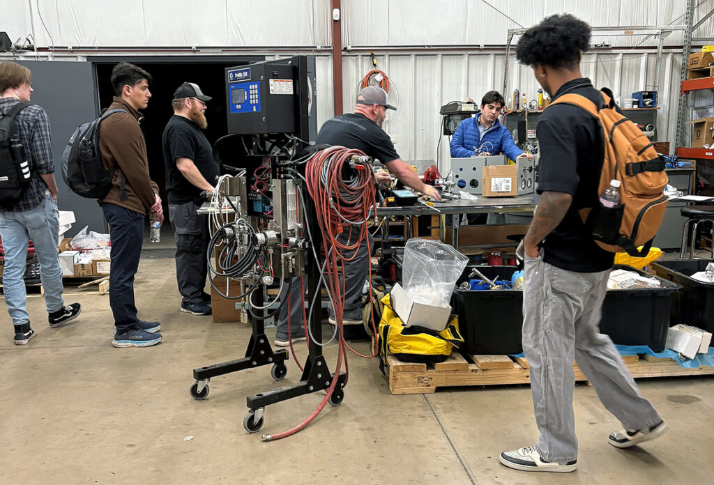 Industrial paint system staging workshop with Bradley University students observing Jason Mikelson of Dove Equipment demonstrating assembly processes at a workbench, supported by mentor Brandon Wolstenholme